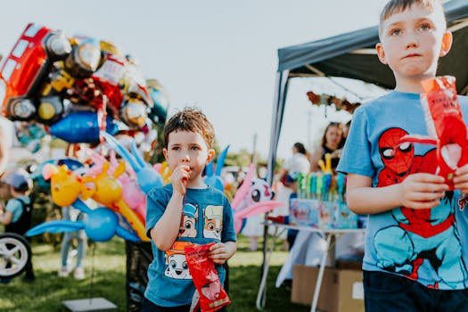 Kids in superhero shirts enjoying candies and colorful balloons at a lively outdoor fair.