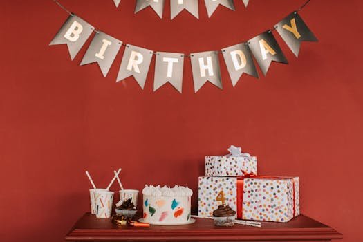 Festive birthday scene with cake, cupcakes, gifts, and decorations against red wall.