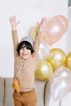 A cheerful child with arms raised surrounded by colorful balloons at an indoor celebration.