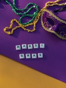 Colorful Mardi Gras mask and beads with Scrabble tiles spelling 'Mardi Gras' on purple surface.