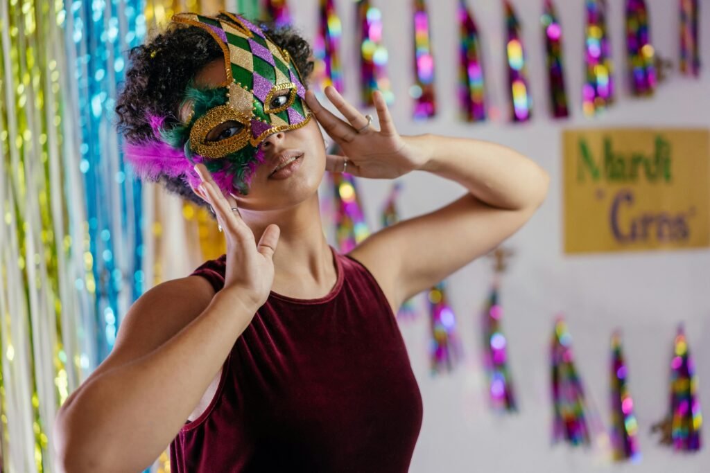 A woman wearing a vibrant Mardi Gras mask poses playfully with colorful decorations in the background.